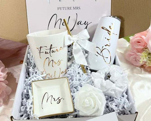 A gift set for bride-to-be including a personalized bridal mug, champagne flute, Mrs. ring dish, and a white satin hair scrunchie, all displayed in an open white gift box with white shred paper.