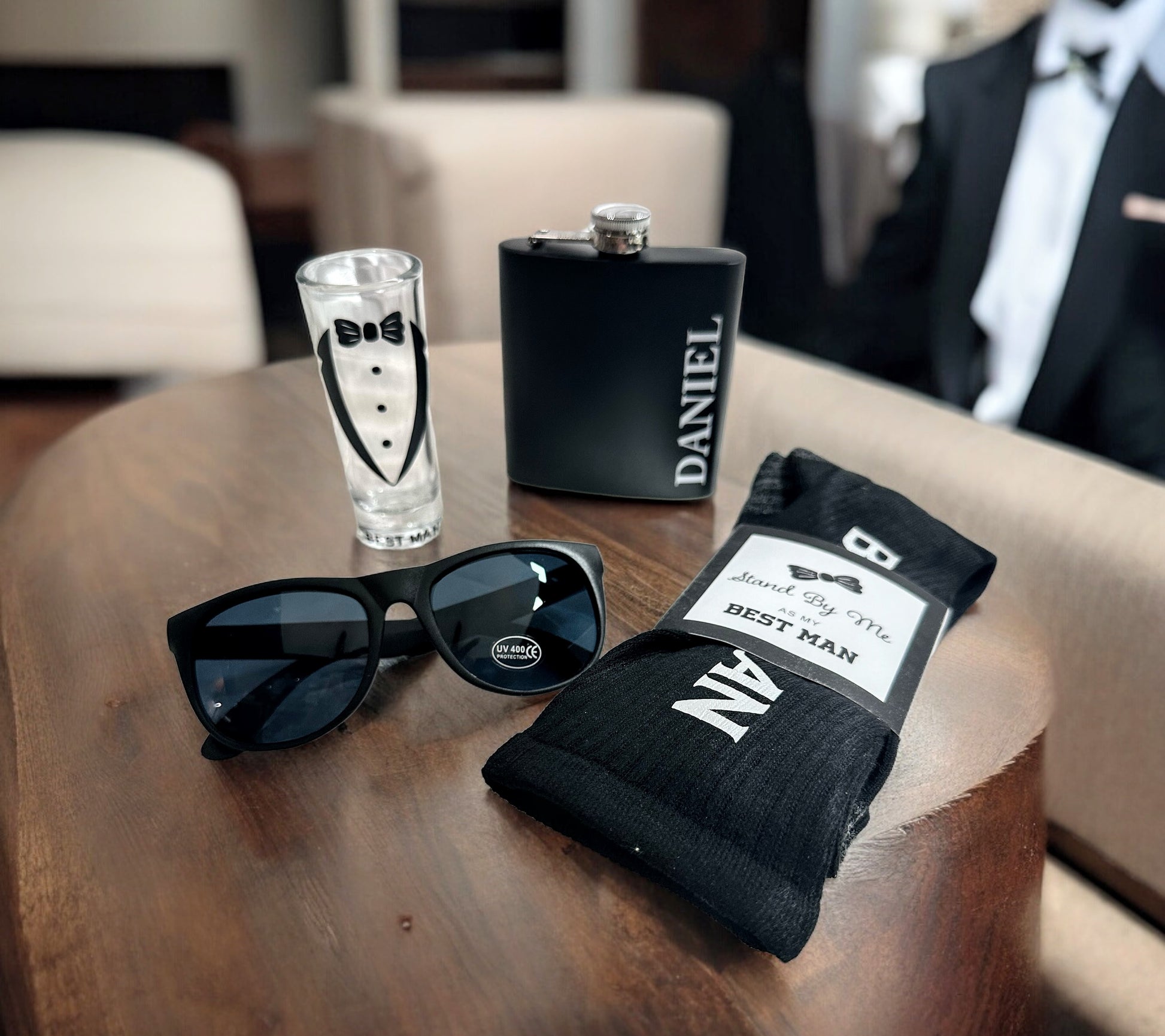 Black sunglasses, shot glass, and black socks with 'Best Man' text on a wooden surface. NIMA Gifts branding.