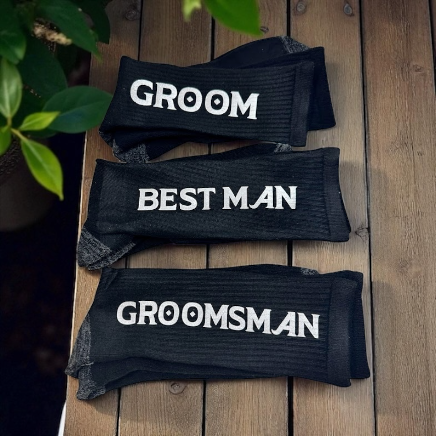 Black socks with 'GROOM', 'BEST MAN', and 'GROOMSMAN' text on a wooden surface.