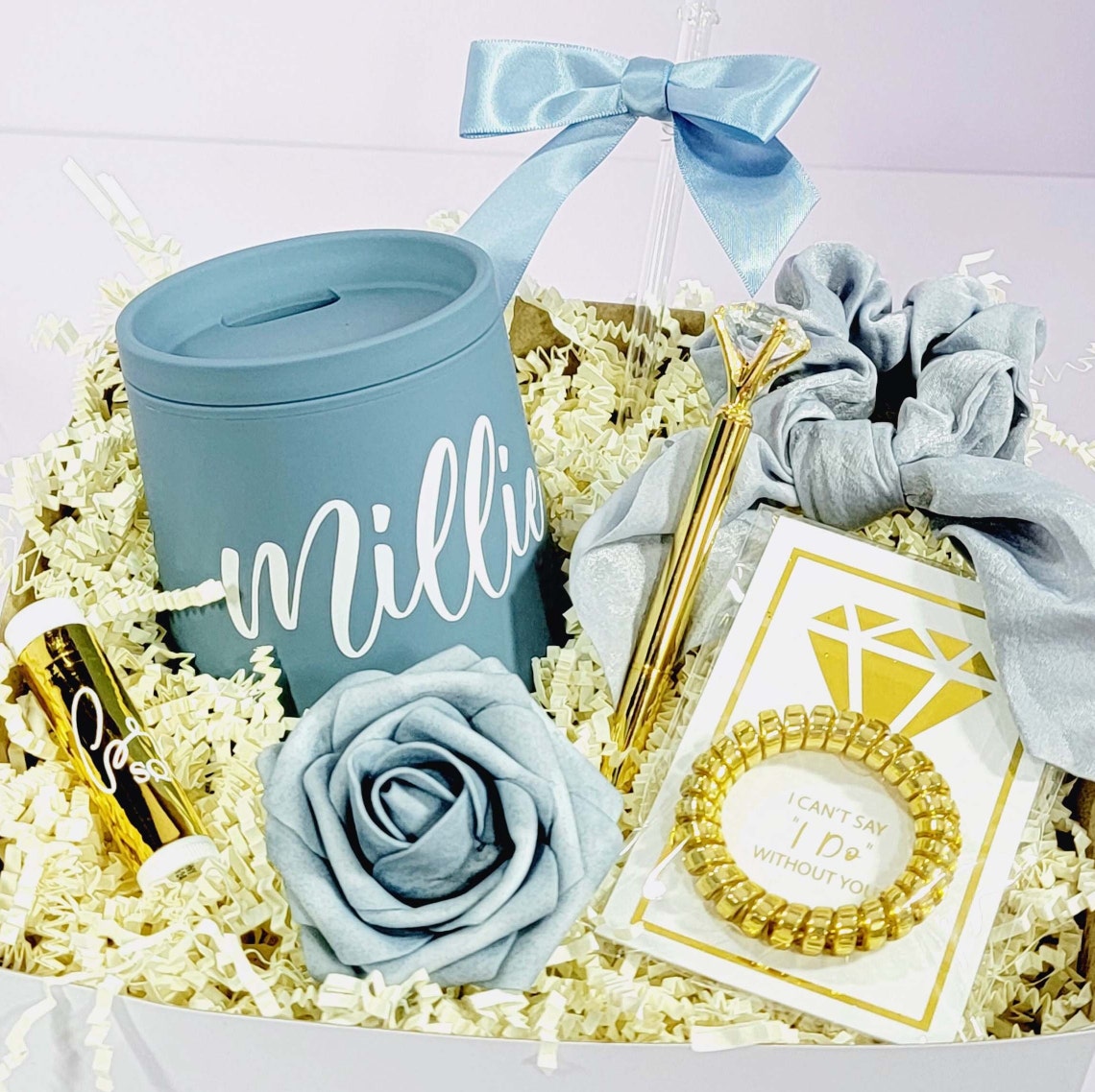 Another variation of the NIMA Gifts Dusty Blue Bridesmaid Proposal Gift Basket which contains a dusty blue tumbler, straw with a dusty blue satin bow, a satin dusty blue bow tie hair scrunchies, gold foiled lip balm, gold telephone wire hair tie on a gold foiled diamond card that says "I Can't Say I Do Without You" and a foam rose to march box color scheme of dusty blue.