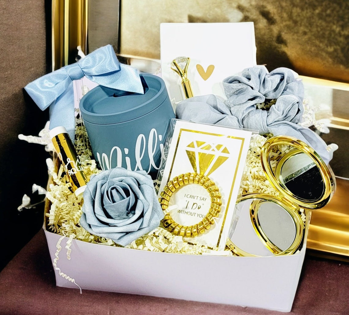 Bridesmaid proposal gift box in Dusty Blue and Gold Wedding Color. The gift box includes a dusty blue tumbler, straw with a dusty blue bow, a gold diamond pen, gold foiled lip balm, "I Can't Say I Do Without You" Gold hair tie on a gold foiled diamond card, personalized gold mirror, dusty blue bow tie hair scrunchy and a foam rose to match gift box colors