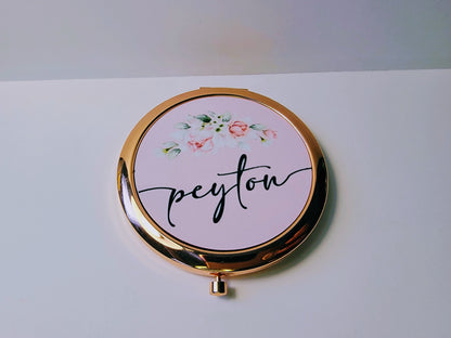 Bridesmaid, jr. bridesmaid, or flower girl Compact mirror personalized with a girls name and with a heart design, placed on a white packaging box with a gold heart, surrounded by flowers. A great thank you gift or a birthday gift for a younger teenager.