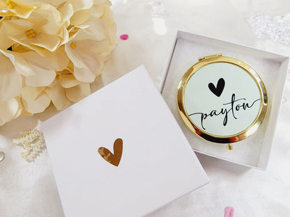 Bridesmaid, jr. bridesmaid, or flower girl Compact mirror personalized with a girls name and with a heart design, placed on a white packaging box with a gold heart, surrounded by flowers. A great thank you gift or a birthday gift for a younger teenager.