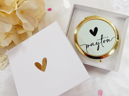 Bridesmaid, jr. bridesmaid, or flower girl Compact mirror personalized with a girls name and with a heart design, placed on a white packaging box with a gold heart, surrounded by flowers. A great thank you gift or a birthday gift for a younger teenager.