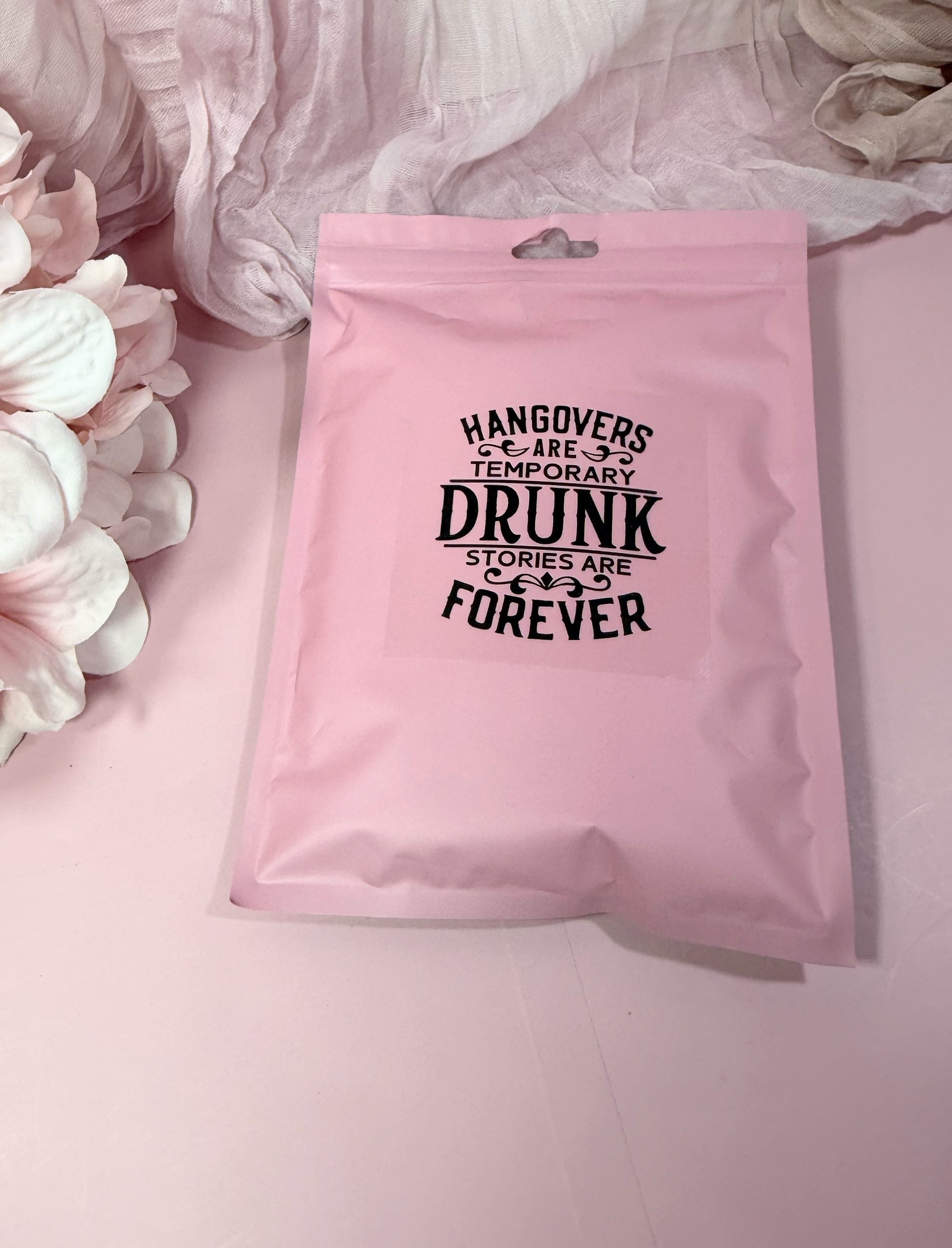 Pink poly pouch package with humorous text on a pink background with flowers showcasing the back side of the product option for the Hangover Recovery Kit by NIMA Gifts