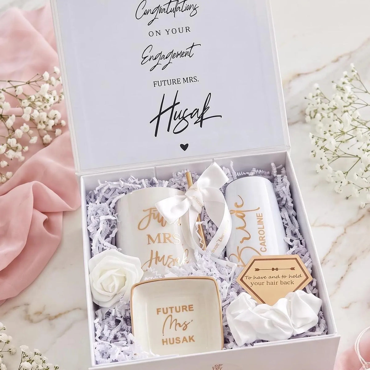 This Personalized Bride to Be Engagement Gift Box is filled with a  personalized coffee mug, personalized bride champagne tumbler with a gold metal straw and personalized satin bow, personalized ring dish, a white satin hair scrunchie and a white foam rose. A greeting card to make your wishes and ask packed in a white magnetic keepsake gift box personalized just for her.