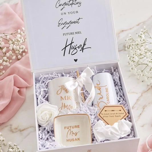 This Personalized Bride to Be Engagement Gift Box is filled with a  personalized coffee mug, personalized bride champagne tumbler with a gold metal straw and personalized satin bow, personalized ring dish, a white satin hair scrunchie and a white foam rose. A greeting card to make your wishes and ask packed in a white magnetic keepsake gift box personalized just for her.