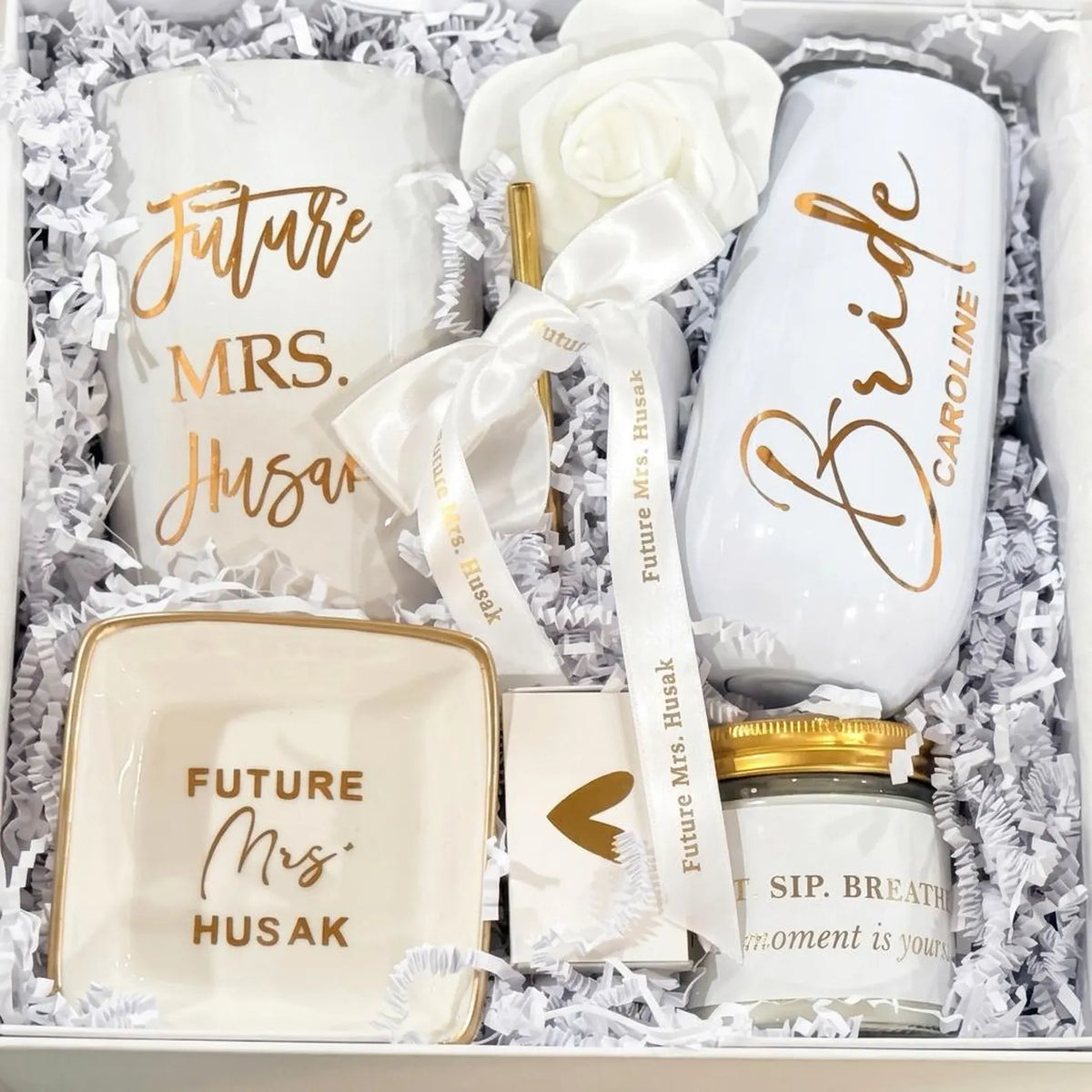 This Personalized Bride to Be Engagement Gift Box is filled with a  personalized coffee mug, personalized bride champagne tumbler with a gold metal straw and personalized satin bow, personalized ring dish, a white satin hair scrunchie and a white foam rose. A greeting card to make your wishes and ask packed in a white magnetic keepsake gift box personalized just for her.