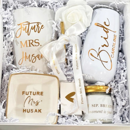 This Personalized Bride to Be Engagement Gift Box is filled with a  personalized coffee mug, personalized bride champagne tumbler with a gold metal straw and personalized satin bow, personalized ring dish, a white satin hair scrunchie and a white foam rose. A greeting card to make your wishes and ask packed in a white magnetic keepsake gift box personalized just for her.