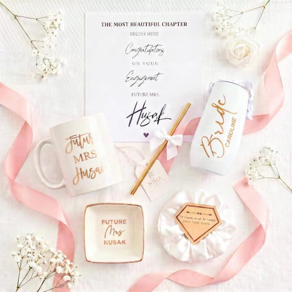 This Personalized Bride to Be Engagement Gift Box is filled with a  personalized coffee mug, personalized bride champagne tumbler with a gold metal straw and personalized satin bow, personalized ring dish, a white satin hair scrunchie and a white foam rose. A greeting card to make your wishes and ask packed in a white magnetic keepsake gift box personalized just for her.