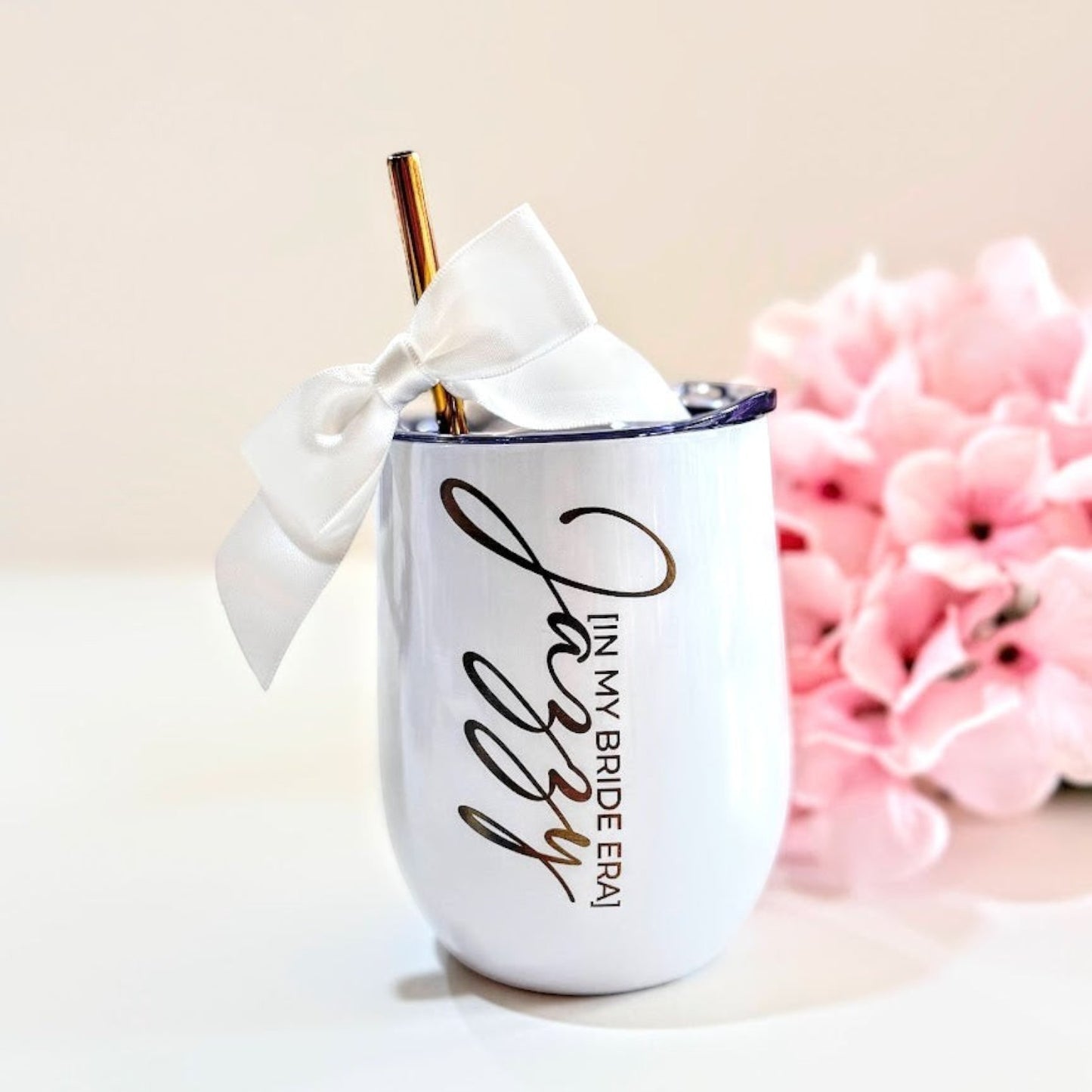 White wine tumbler with gold text and straw, tied with a white ribbon, against a light background with pink flowers.