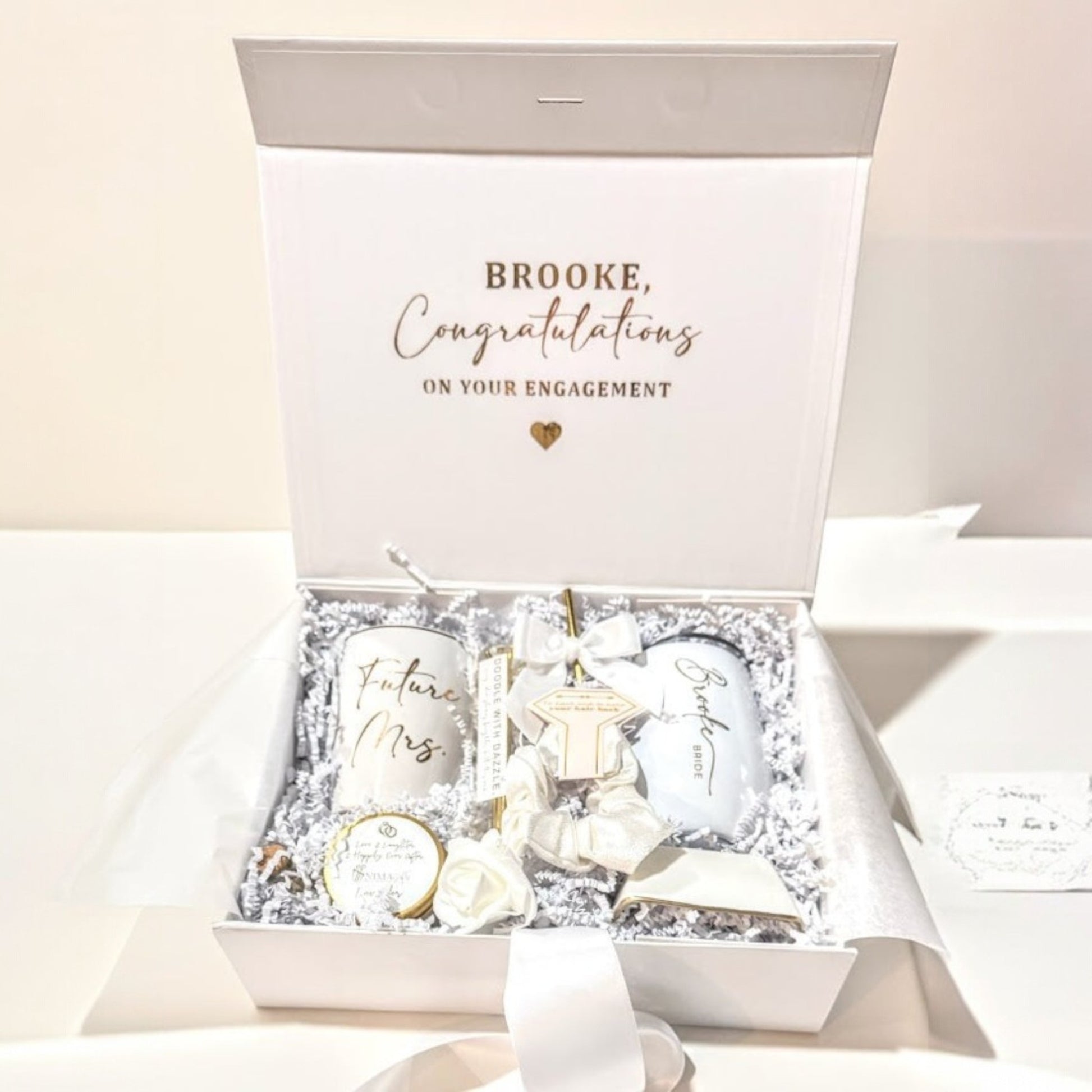 A just engaged Gift set for a bride to be engagement  gift set that shows all the items that are included which are a personalized coffee mug, and personalized engraved wine tumbler, a white gold bling pen, an initialed porcelain ring dish with gold rim, a hand poured lavender candle with gold label and a white satin ribbon with a white satin bow, a  sparkle and a rose on the candle and a bottle of matches with a rose and showing a personalized keepsake magnetic gift box with the bride to be name on it.