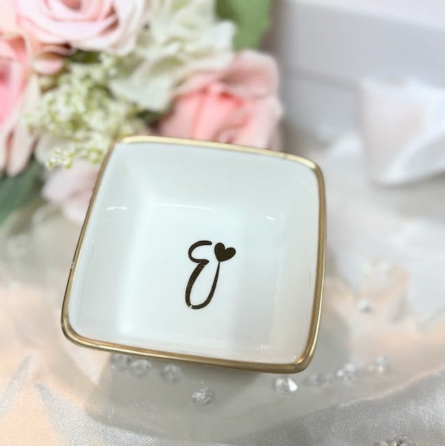 A square personalized ring dish with a decorative letter 'E' and gold rim is sitting on a table with flowers and jewelry.