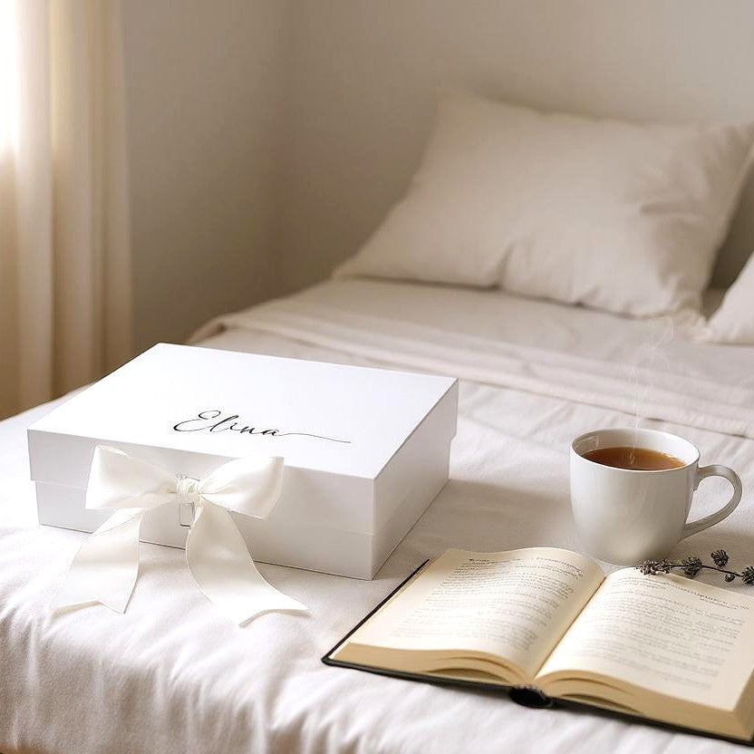 A white engagement keepsake gift box with a gold name on the box with a white satin bow tied neatly on the front of the box, an open book, and cup of coffee on a bed in a softly lit room.