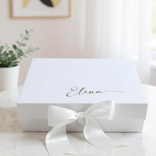 A white engagement keepsake magnetic gift box with a gold name on top and a a white satin bow neatly tied in front of the box, is sitting on a marble table.