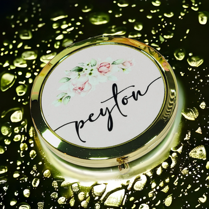 Bridesmaid, jr. bridesmaid, or flower girl Compact mirror personalized with a girls name and with a heart design, placed on a white packaging box with a gold heart, surrounded by flowers. A great thank you gift or a birthday gift for a younger teenager.
