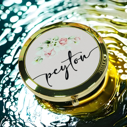 Bridesmaid, jr. bridesmaid, or flower girl Compact mirror personalized with a girls name and with a heart design, placed on a white packaging box with a gold heart, surrounded by flowers. A great thank you gift or a birthday gift for a younger teenager.