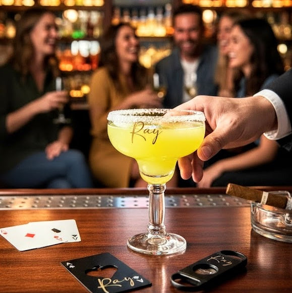 A man is about to be holding a personalized margarita glass with the name 'Ray' on the front of the glass, the glass is on a bar counter with a personalized beer bottle opener and with a cigar cutter by the glass. In the background , there's a group of men and women talking and laughing seemingly celebrating an event.