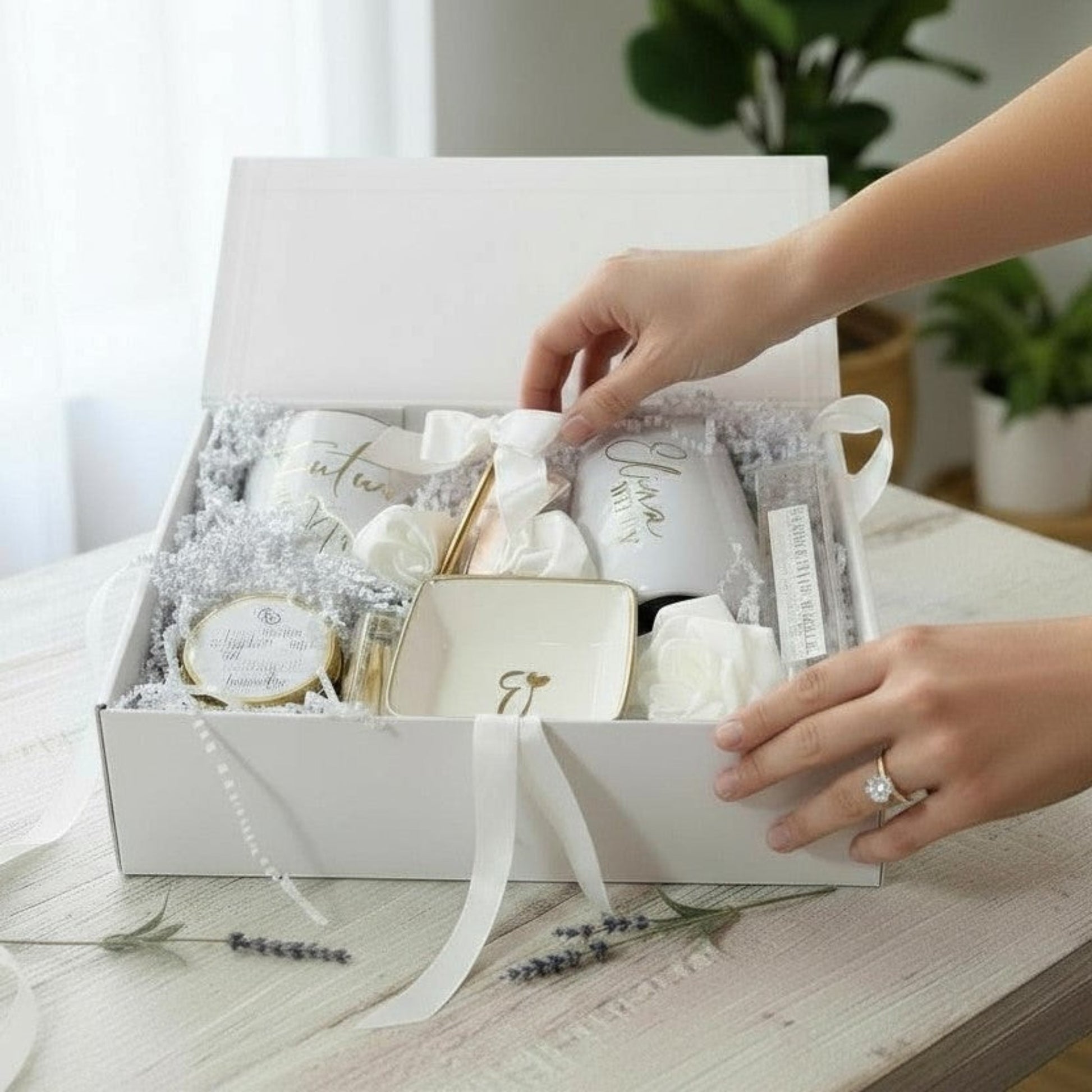 Person opening a white engagement gift box at a bridal shower which is filled with a personalized mug that says Future Mrs on it, an engraved and personalized wine tumbler tumbler that says either Bride, Wifey or In My Bride Era with a gold straw and a satin bow, a ring dish with gold rim and an initial, a hand poured candle with matches and a bling pen. All finished with a white bow, in a white keepsake magnetic engagement gift box.