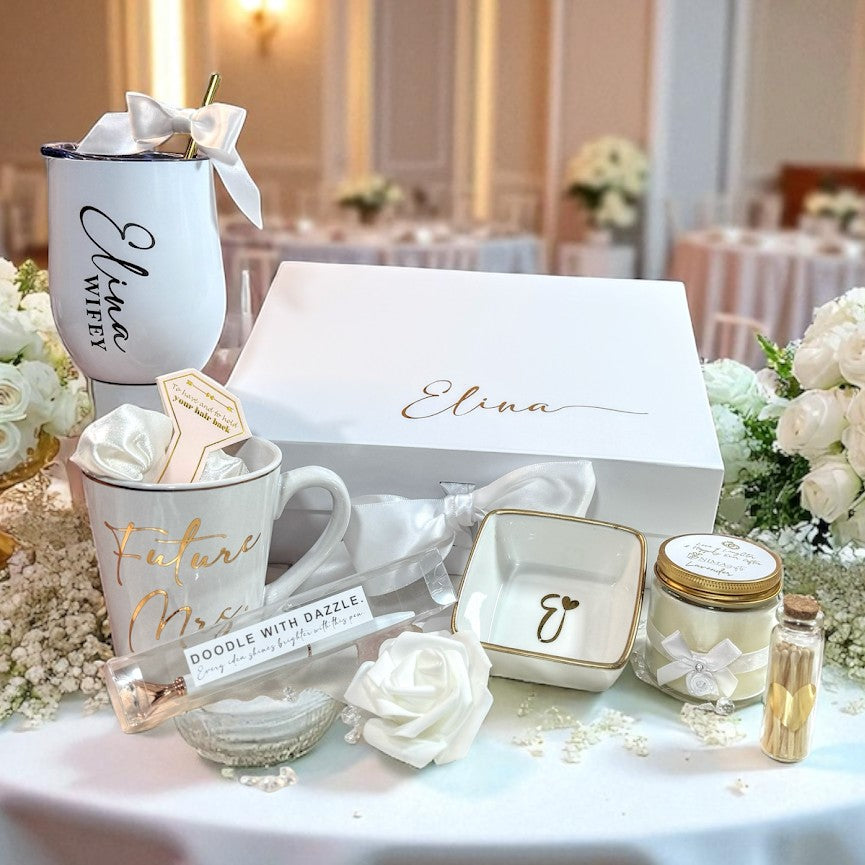 Table setting  displaying a bride to be engagement  gift set that shows all the items that are included which are a personalized coffee mug, and personalized engraved wine tumbler, a white gold bling pen, an initialed porcelain ring dish with gold rim, a hand poured lavender candle with gold label and a white satin ribbon with a white satin bow, a  sparkle and a rose on the candle and a bottle of matches with a foam rose and showing a personalized keepsake magnetic gift box with the bride to be name on it.
