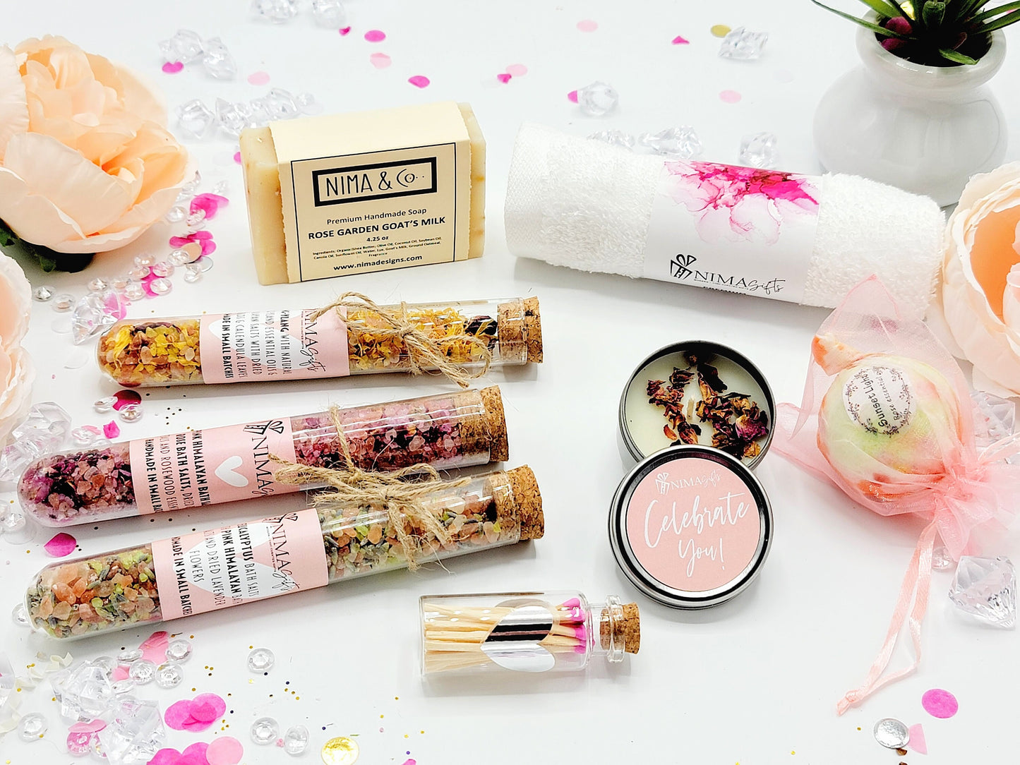 A spa gift set with a pink and white theme. The set includes a bar of soap, a bath towel, a bath bomb, a candle, a small bottle of matches, and three glass tubes filled with bath salts. The bath bomb is wrapped in pink tulle and has a pink and white floral design. The candle is in a small metal tin with a pink lid and the words 'Celebrate You!' printed on it. The matches are in a small glass bottle with a cork top. The bath salts are in glass tubes and complete the gift box.