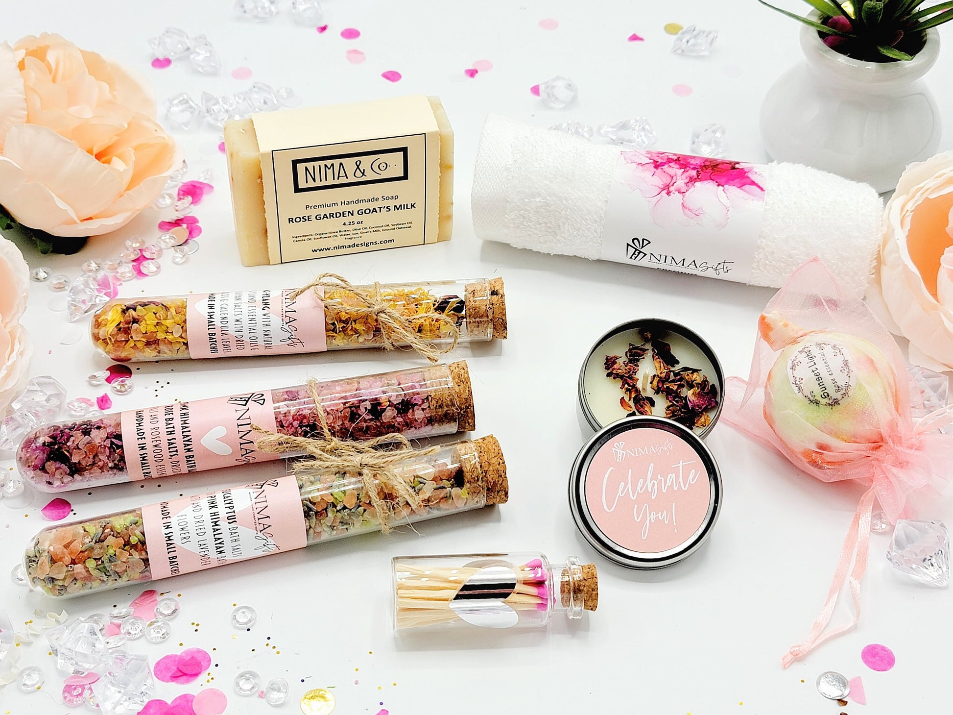 A spa gift set with a pink and white theme. The set includes a bar of soap, a bath towel, a bath bomb, a candle, a small bottle of matches, and three glass tubes filled with bath salts. The bath bomb is wrapped in pink tulle and has a pink and white floral design. The candle is in a small metal tin with a pink lid and the words 'Celebrate You!' printed on it. The matches are in a small glass bottle with a cork top. The bath salts are in glass tubes and complete the gift box.