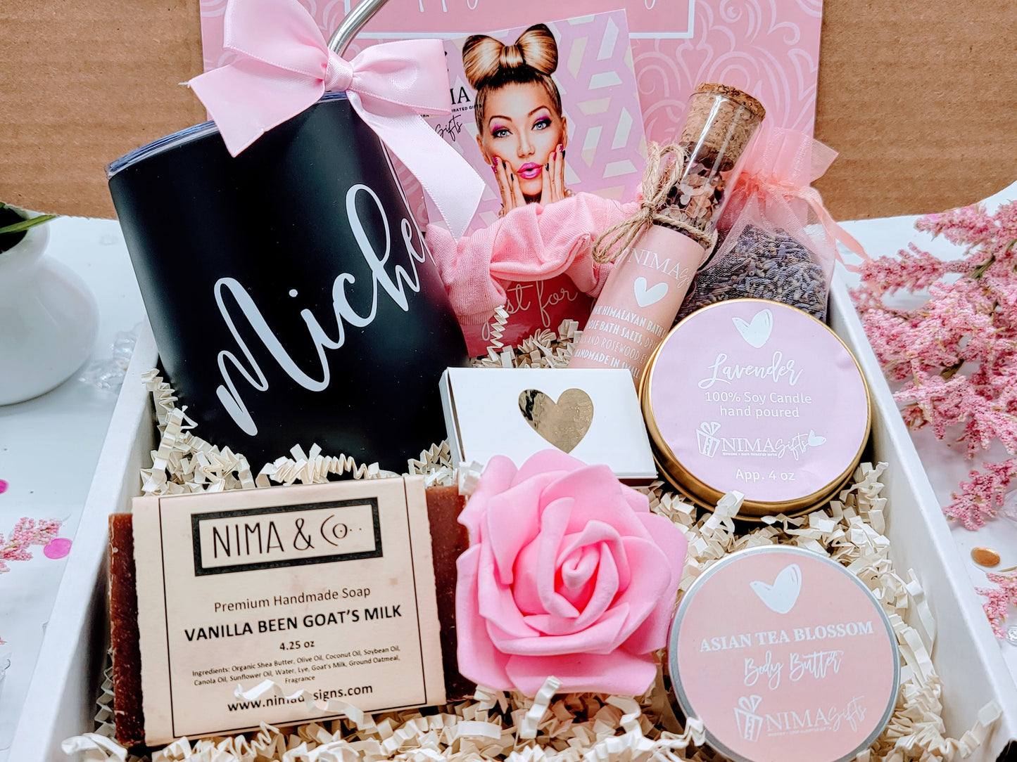 Open view of personalized spa gift box showing a colored wine tumbler, metal straw with satin bow, lavender soy candle with dried flowers, glass bath crystal tube, printed greeting card, foam rose, and shredded paper inside a custom-design box.
