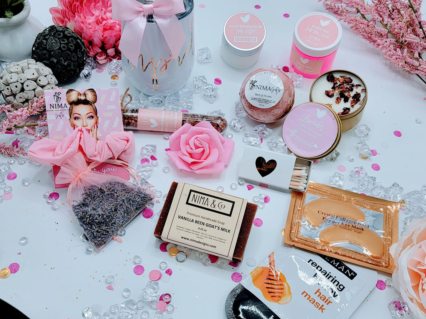 Top-down shot of personalized spa gift box with custom-name lid, showing a colored wine tumbler, metal straw with satin bow, lavender soy candle with dried flowers, glass bath crystal tube, printed greeting card, foam rose, and shredded paper inside.