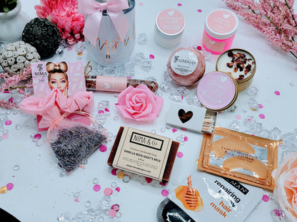 Top-down shot of personalized spa gift box with custom-name lid, showing a colored wine tumbler, metal straw with satin bow, lavender soy candle with dried flowers, glass bath crystal tube, printed greeting card, foam rose, and shredded paper inside.