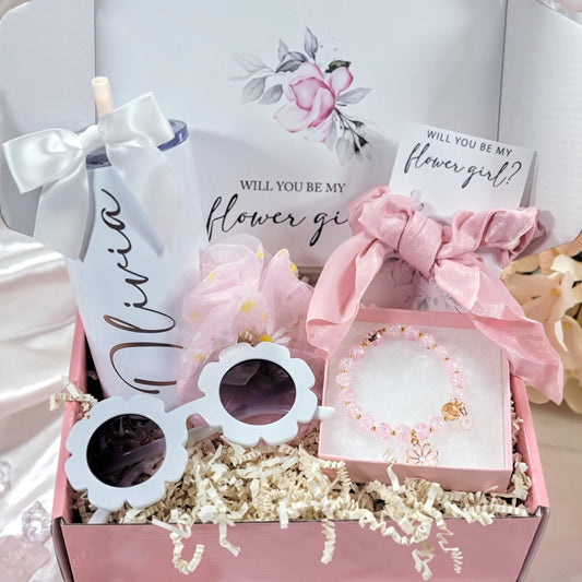 A pink gift box filled with items for a flower girl. The box contains a white tumbler with a straw and the name Olivia on it, a pink hair scrunchie, white sunglasses with a daisy shape, a pink bracelet with a daisy charm, and a pink card that says 'Will you be my flower girl?'