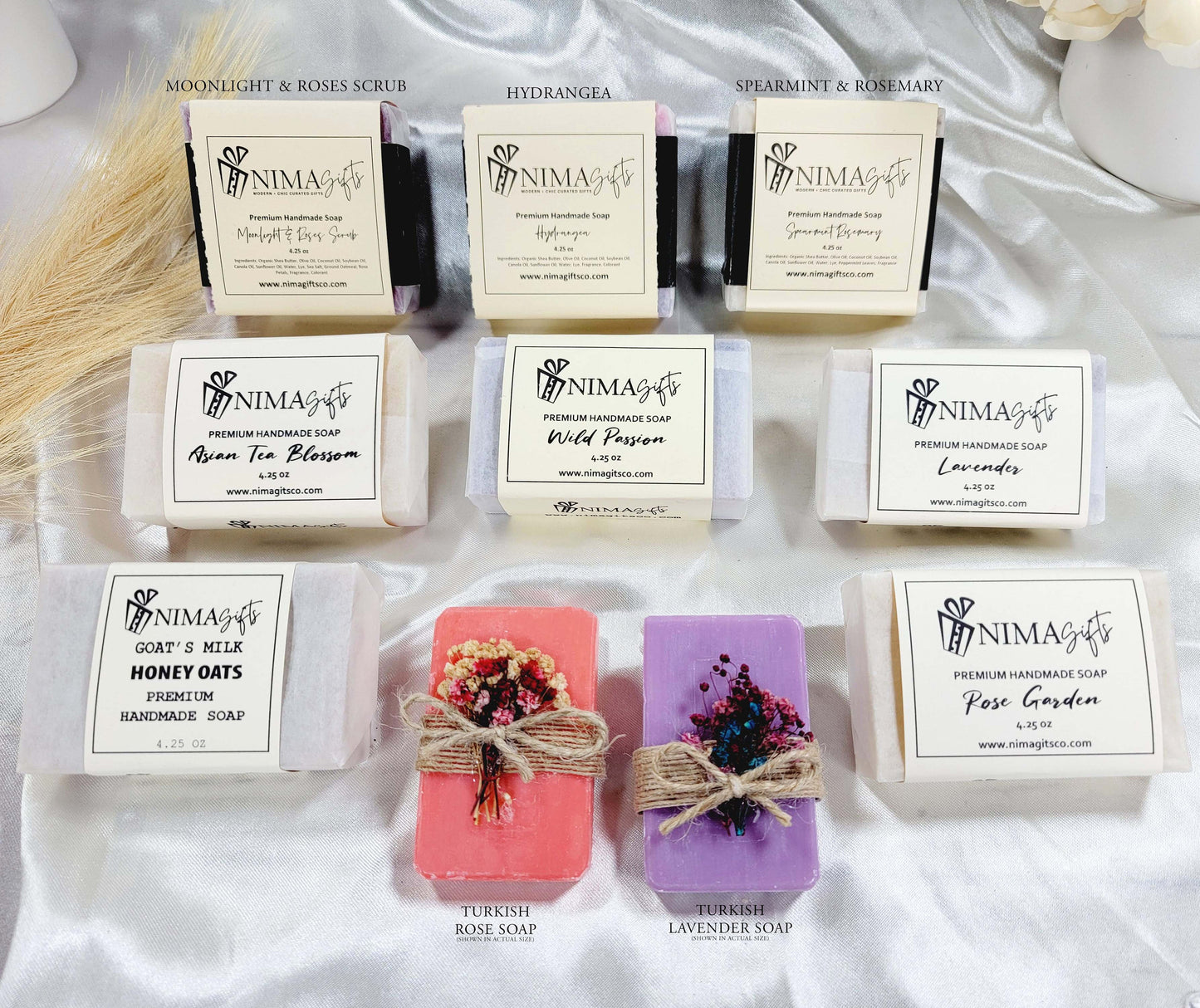 Nine handmade soap bars in assorted scents and colors, each wrapped in white label paper with scent name and NIMA Gifts branding, arranged in three rows including floral, herbal, and exfoliating varieties.