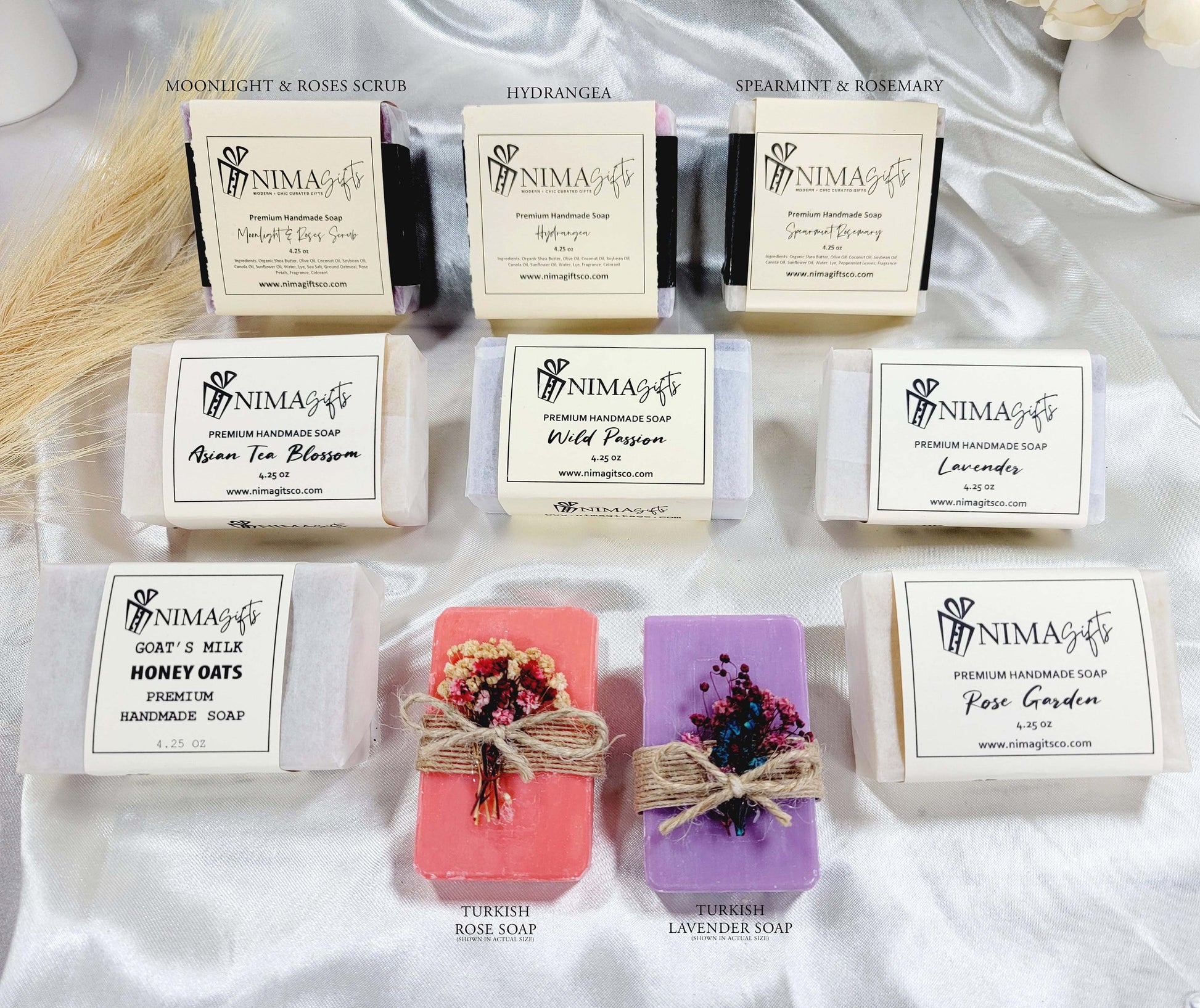 Nine handmade soap bars in assorted scents and colors, each wrapped in white label paper with scent name and NIMA Gifts branding, arranged in three rows including floral, herbal, and exfoliating varieties.