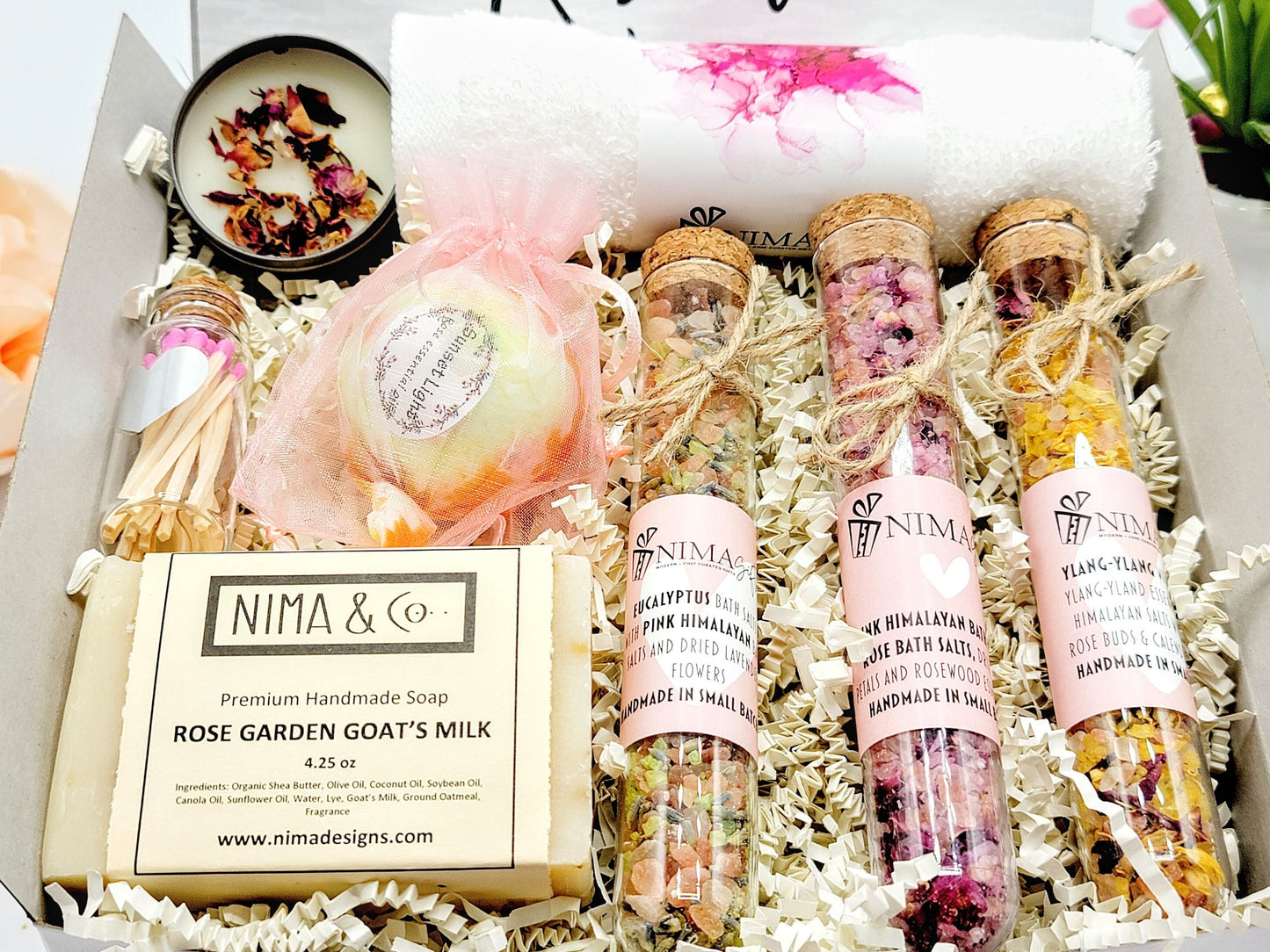 A gift box filled with bath and body products. The gift contains a white wash towel, a small bath bomb, three glass tubes filled with bath salts, and a Premium Handmade Soap, and a small candle with matches to finish the gift box.