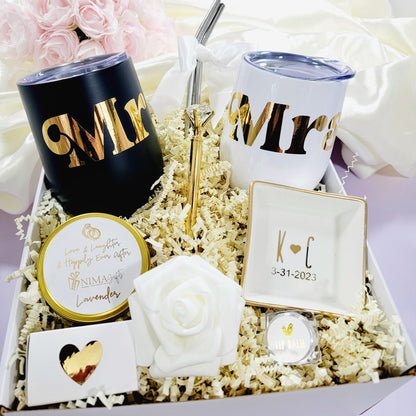 This Couples Engagement Gift Box includes a white gift box filled with wedding-themed gifts, including a black and white insulated tumblers with gold lettering that reads "Mr." and "Mrs.", a small white dish with gold trim and the initials "K-C" and the EST date "3-31-2023", a small white rose, a candle with a gold ring design and the text "Love & Laughter & Happily Ever After", and a small container of lip balm.