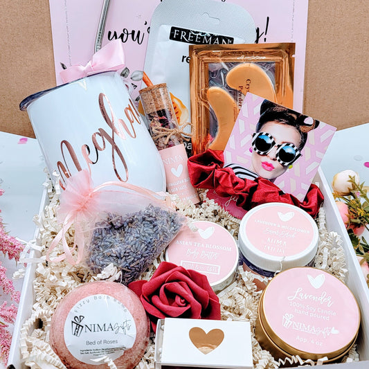Open view of personalized spa gift box showing a colored wine tumbler, metal straw with satin bow, lavender soy candle with dried flowers, glass bath crystal tube, printed greeting card, foam rose, and shredded paper inside a custom-design box.”