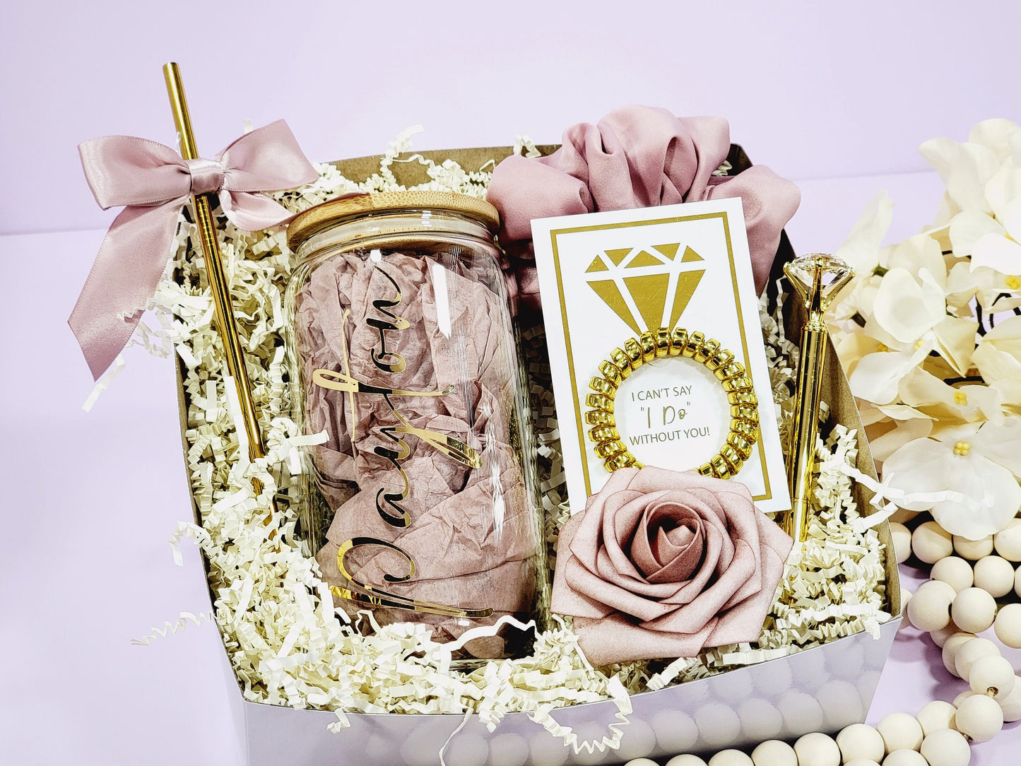 A personaluzed bridesmaid proposal gift box for Future Mrs or Bride to Be, includes a gold pen, a compact mirror, a hair tie, and a card with a gold diamond graphic. The box also contains a personalized glass with a name on it, a bow, and a decorative rose. The color scheme is primarily dusty rose and gold.