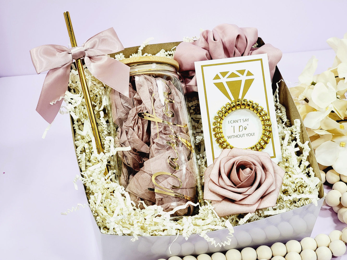 A bridesmaid proposal gift box with a wedding theme. It contains a personalized glass jar with a bamboo lid,  with a gold straw and bow, a card with a ring design and the text "I can't say I do without you!", a scrunchie, a rose, and decorative paper.