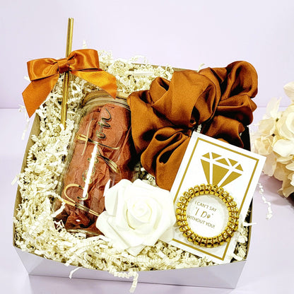 A bridesmaid proposal gift box that contains a 16oz glass tumbler with gold lettered name, gold straw with matching satin bow, a satin burnt orange hair scrunchie, a gold telephone wire scrunchie by NIMA Gifys