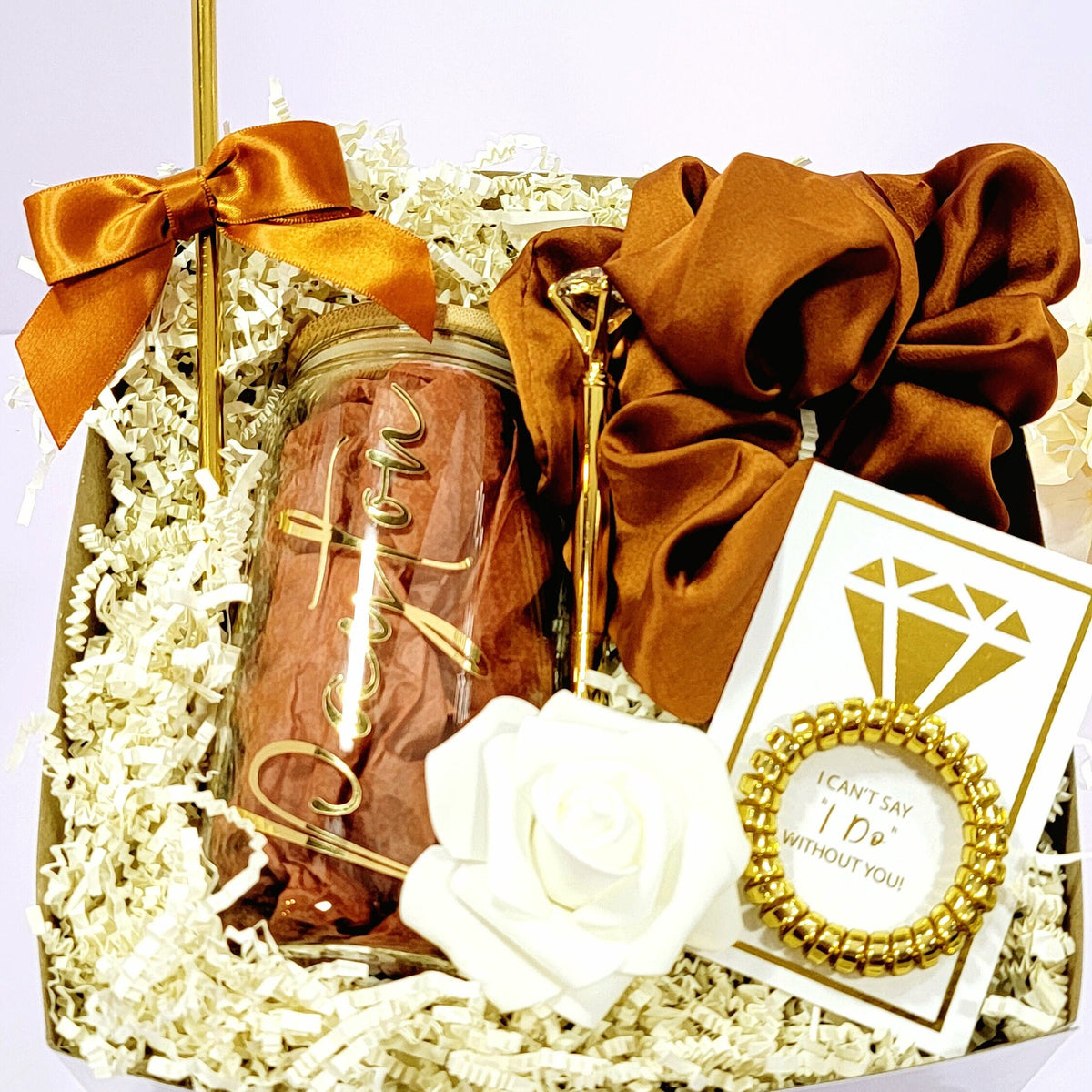 A bridesmaid proposal gift box that contains a 16oz glass tumbler with gold lettered name, a gold diamond pen, gold straw with matching satin bow, a satin burnt orange hair scrunchie, a gold telephone wire scrunchie by NIMA Gifts