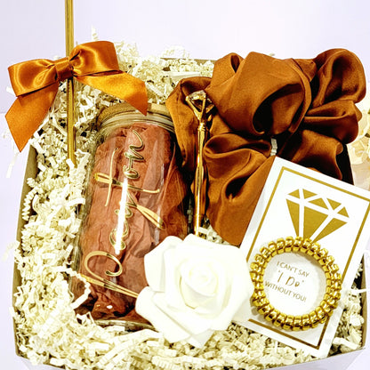 A bridesmaid proposal gift box that contains a 16oz glass tumbler with gold lettered name, a gold diamond pen, gold straw with matching satin bow, a satin burnt orange hair scrunchie, a gold telephone wire scrunchie by NIMA Gifts