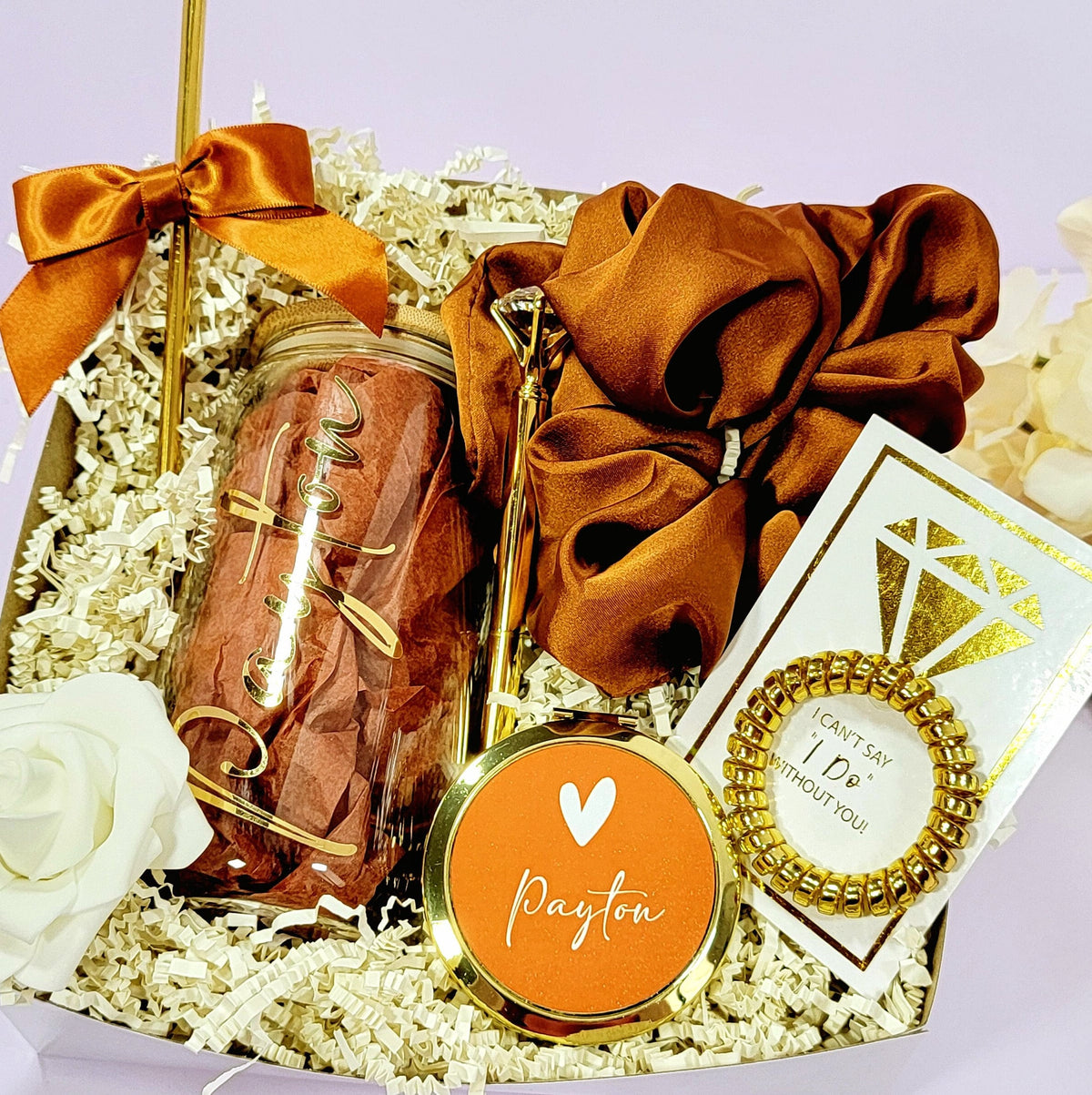 A bridesmaid proposal gift box that contains a 16oz glass tumbler with Clear glass tumbler with gold lettered name, a gold diamond pen, gold straw with matching satin bow, a satin burnt orange hair scrunchie, a gold telephone wire scrunchie and a personalized gold compact mirror by are all put together in a hard top white bix which is personalized on top with the girls name and inside with “Will you be my…Bridesmaid or Main of Honor or Matron of Honor”