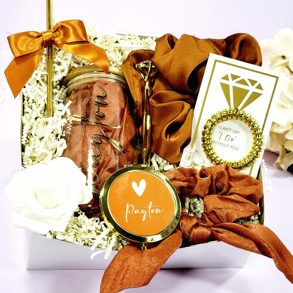 A bridesmaid proposal gift box that contains 16oz glass tumbler with gold lettered name, a gold diamond pen, gold straw with matching satin bow, a satin burnt orange hair scrunchie, a gold wire scrunchie and a gold compact mirror & hair tie scrunchie