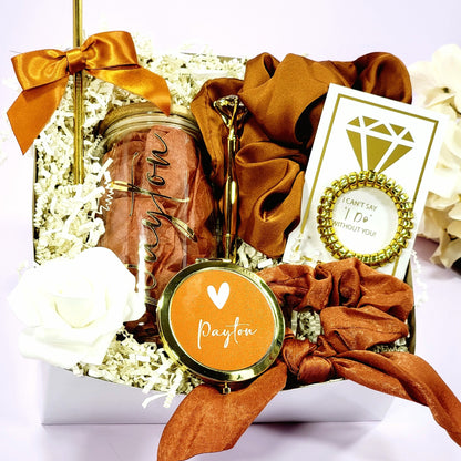 A bridesmaid proposal gift box that contains 16oz glass tumbler with gold lettered name, a gold diamond pen, gold straw with matching satin bow, a satin burnt orange hair scrunchie, a gold wire scrunchie and a gold compact mirror & hair tie scrunchie