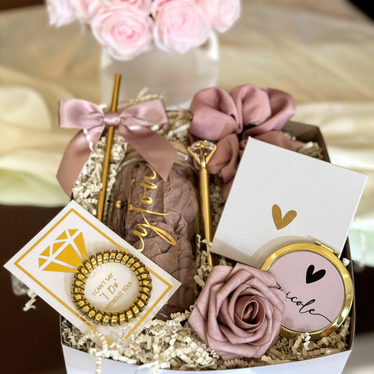 A personaluzed bridesmaid proposal gift box for Future Mrs or Bride to Be, includes a gold pen, a compact mirror, a hair tie, and a card with a gold diamond graphic. The box also contains a personalized glass with a name on it, a bow, and a decorative rose. The color scheme is primarily dusty rose and gold.