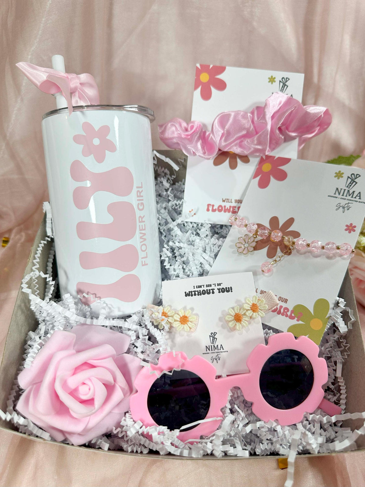 A personalized flower girl proposal gift box that includes a personalized tumbler, a pink hair scrunchie, a pink flower bracelet, a knitted set of flower hair clips, flower girl sunglasses and a pink foam rose by NIMA Gifts
