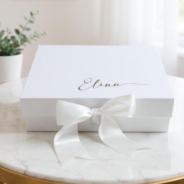 A rectangular white magnetic gift box featuring the name Elina written in gold cursive script on the lid. The box is secured with a wide, white satin ribbon tied into a large bow at the front center, creating a clean and simple presentation.