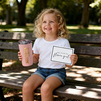 Little girl holding a stainless steel blush tumbler personalized with her name and the words "Flower Girl" on the tumbler in gold letters. She is also holding a card that says: "Will you be our Flower Girl?" This is a part of a larger flower girl proposal gift box that contains Flower Girl Proposal Gift Bag with PU leather handles, filled with personalized keepsakes like tumbler, scrunchie, sunglasses, and greeting card.