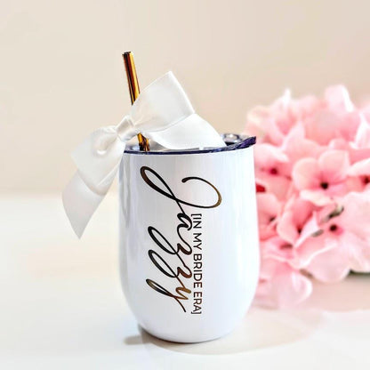 A white tumbler featuring the gold text "Jazzy [IN MY BRIDE ERA]" in a script and sans-serif font. The tumbler includes a gold metal straw adorned with a white satin ribbon tied in a bow at the top, creating a decorative accent.
