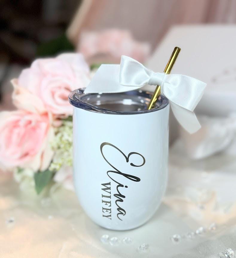 A white stainless steel tumbler with a clear lid and a gold metal straw. The tumbler features the black text Elina Wifey printed vertically on the side. A white satin ribbon bow is tied around the top of the gold metal straw.