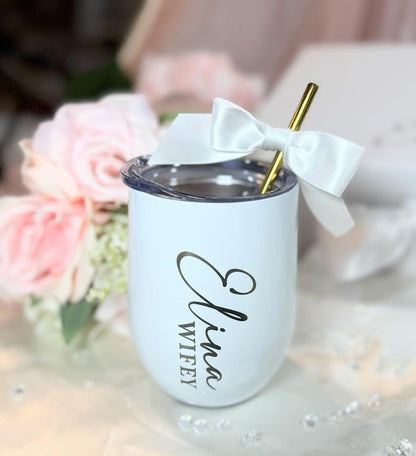 A white stainless steel tumbler with a clear lid and a gold metal straw. The tumbler features the black text Elina Wifey printed vertically on the side. A white satin ribbon bow is tied around the top of the gold metal straw.
